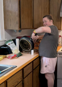 A developmentally disabled young woman with her hair clipped back off her face, is wearing a gray t-shirt and pink athletic shorts, as she scrapes food out of a large frying pan.