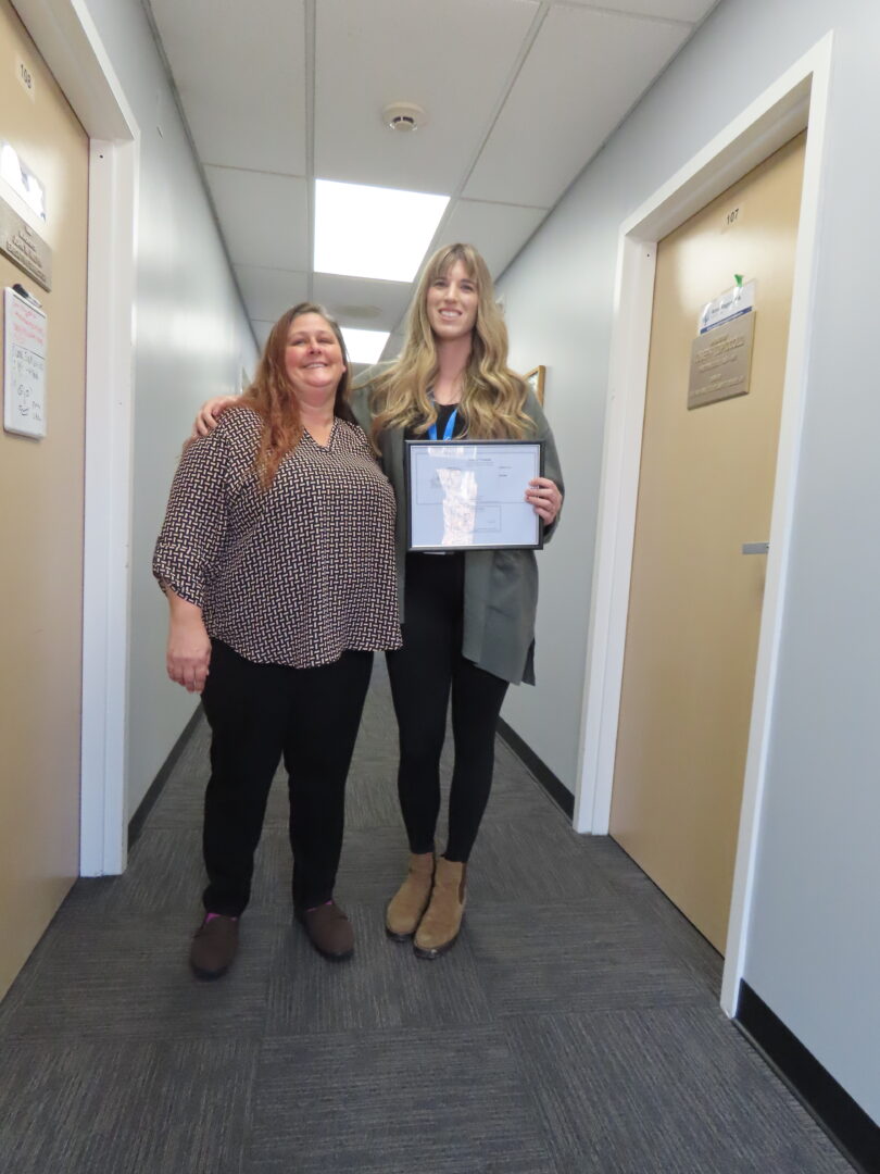 Two smiling women stand side by side in a hallway. Woman on the right with long blonde hair wearing a long gray sweater holds a certificate.
