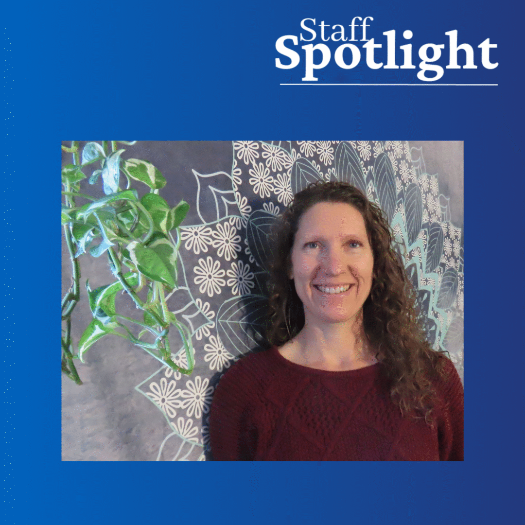 a women with long curly hair and a red sweater smiles against a blue tapestry background with a cascading green plant in the left foreground. Staff Spotlight at the top right of the image