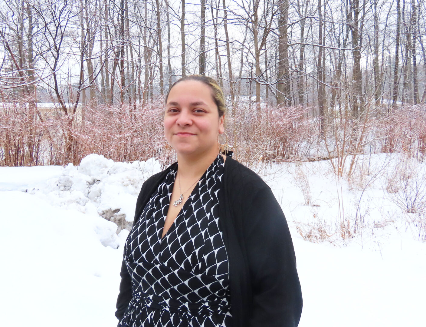 a woman with brunette and blonde hair wearing a black and white shirt with a black sweater stands against a snowy background with bare trees.