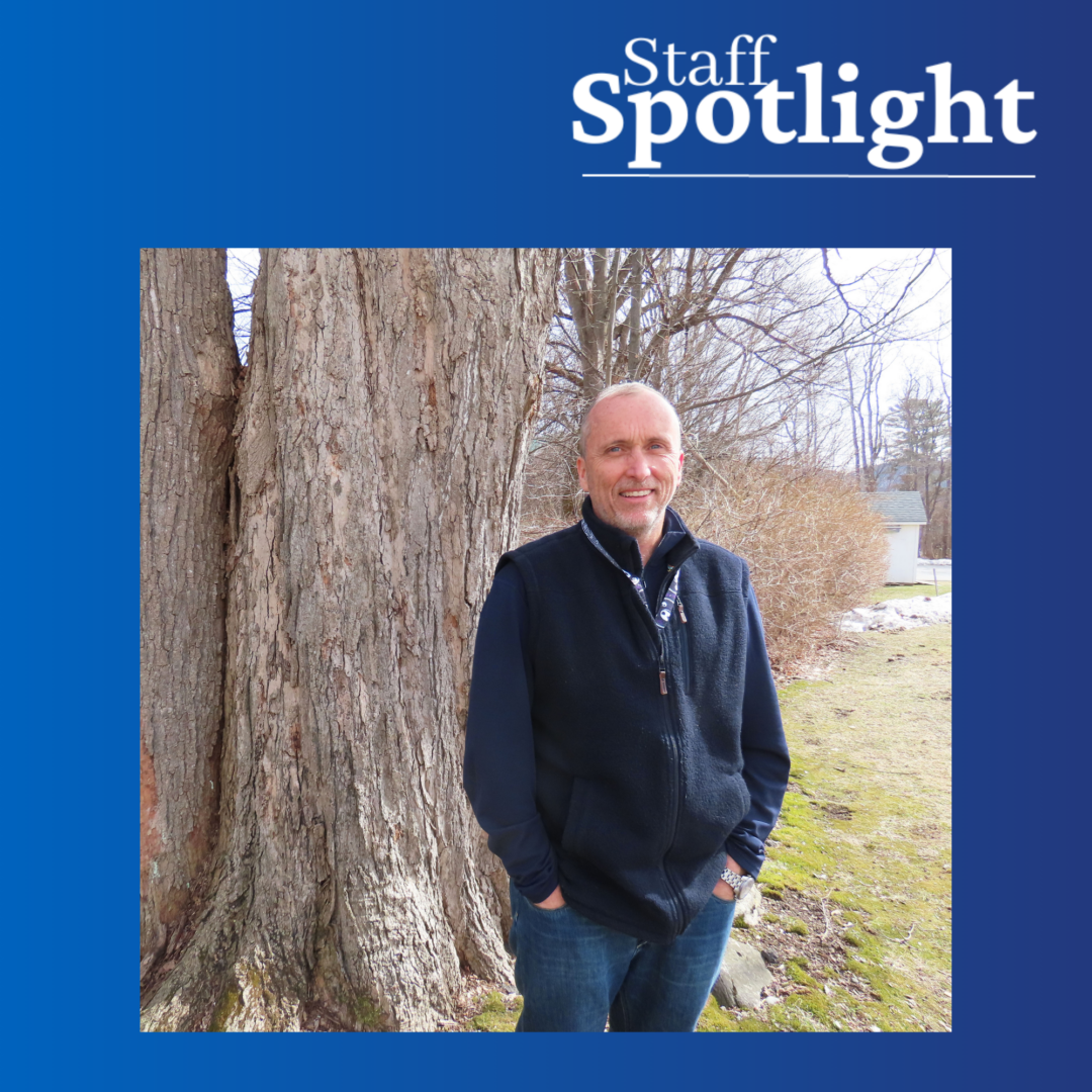 Photo of a man with a dark colored vest and shirt and jeans smiling with a tree and grass in the background. Staff Spotlight in top right with blue gradient background