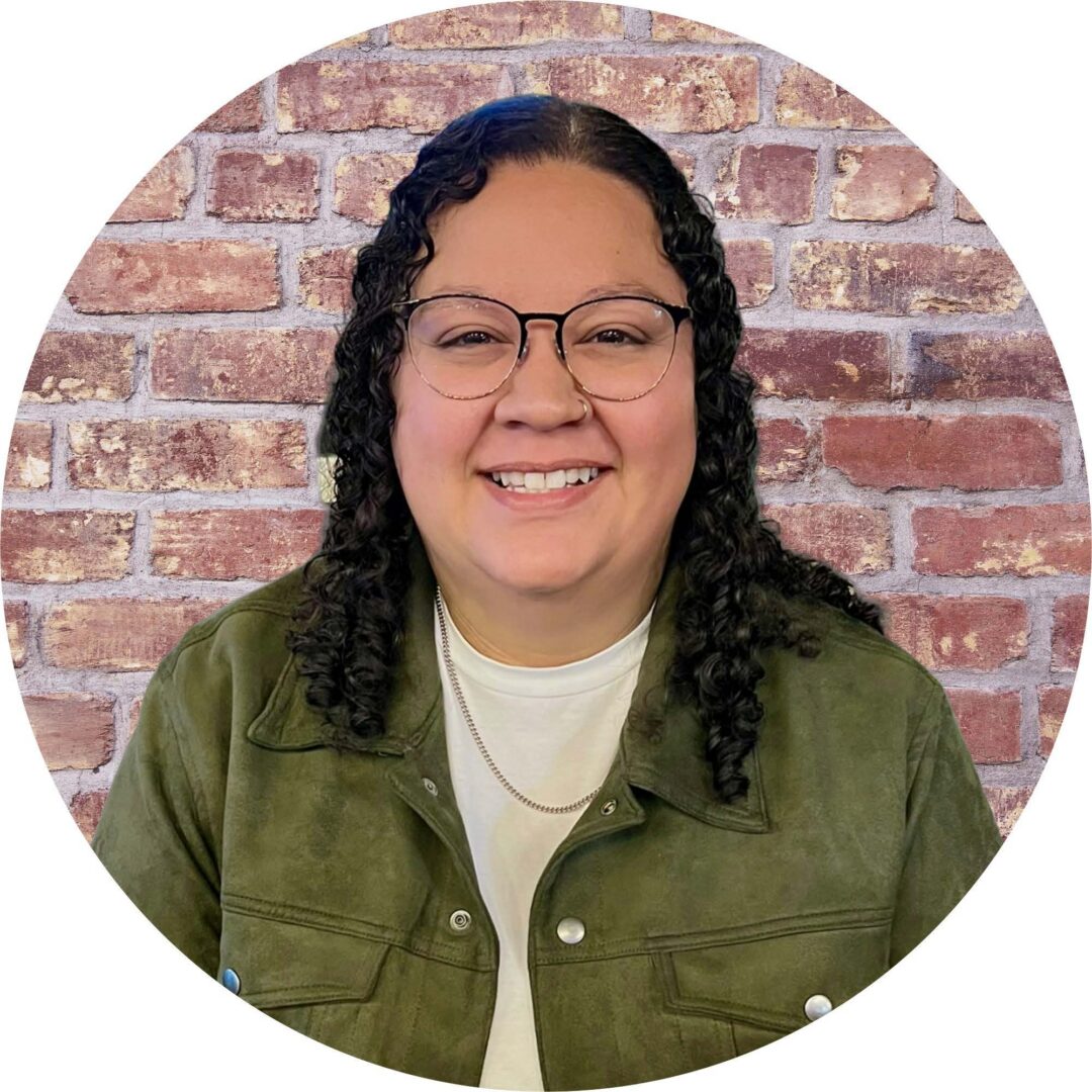 woman with long curly black hair, glasses, a gold chain necklace, white shirt, and green jacket smiles against a brick background.