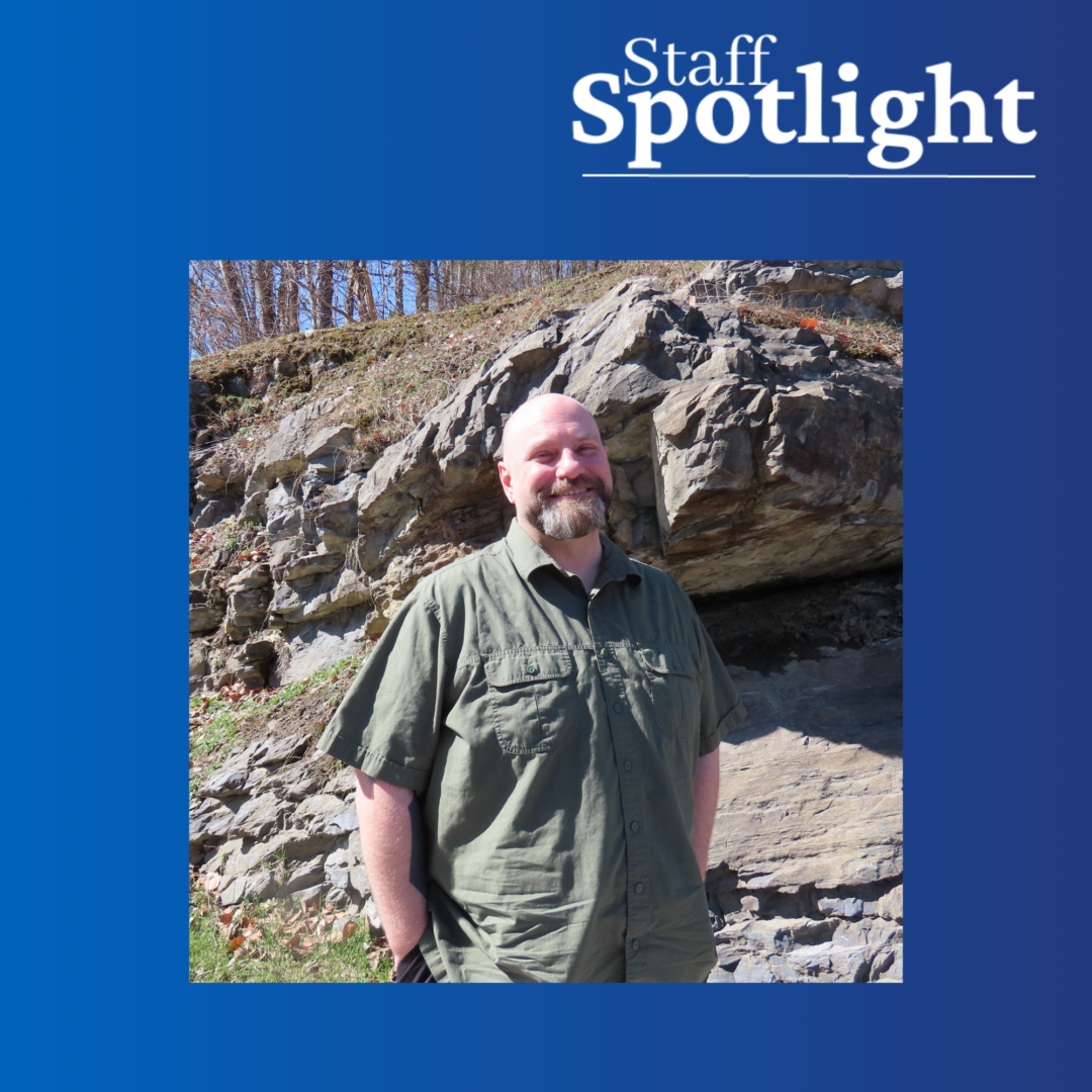 Man with a beard and wearing a green short sleeved button down shirts smiles in front of a rocky outcropping on a sunny day. Staff Spotlight text on top right with a blue gradient border around the whole image.