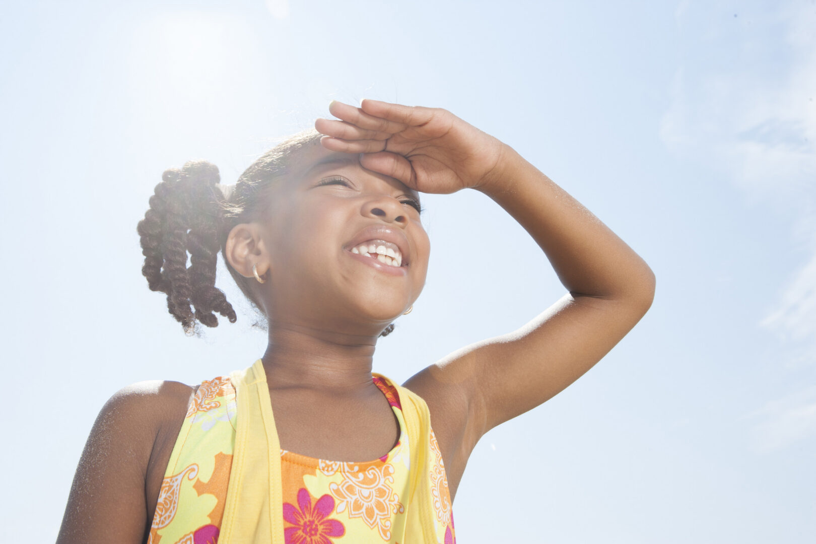 Black girl smiling and looking up at sky with a hand blocking the sun out of her eyes