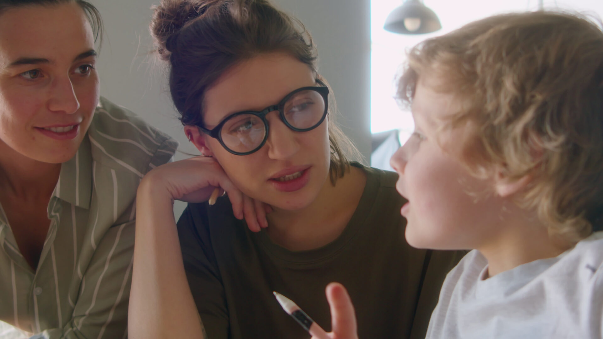 Little caucasian boy holding pen and talking to two women doing homework