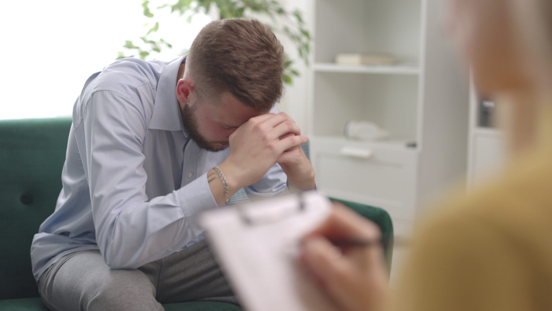 Doctor making notes during a therapy session with a man patient with his head in his hands