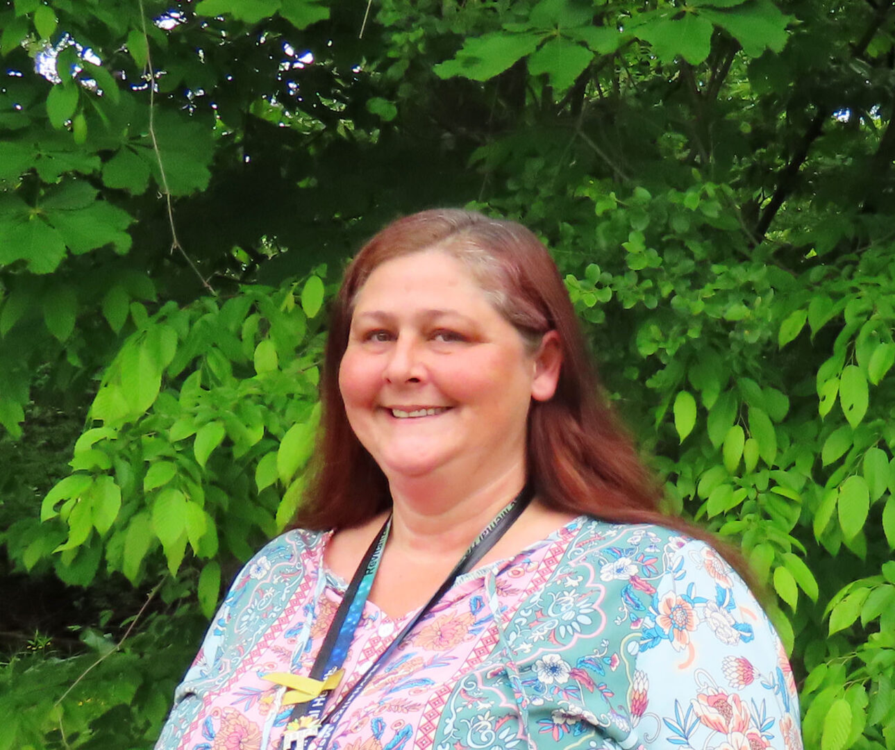 woman with long reddish brunette hair and wearing a floral shirt and blue lanyard smiles against a green leafy background.