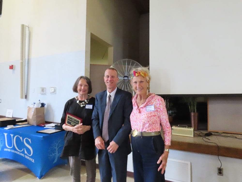two older women smile with a man in a blue suit standing between them in a large room