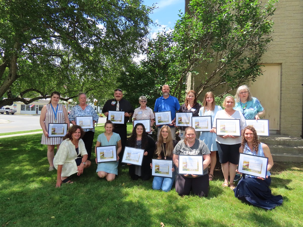 A group of people smile in front of a tan building with a blue sky, green grass and trees in the background while holding certificates.