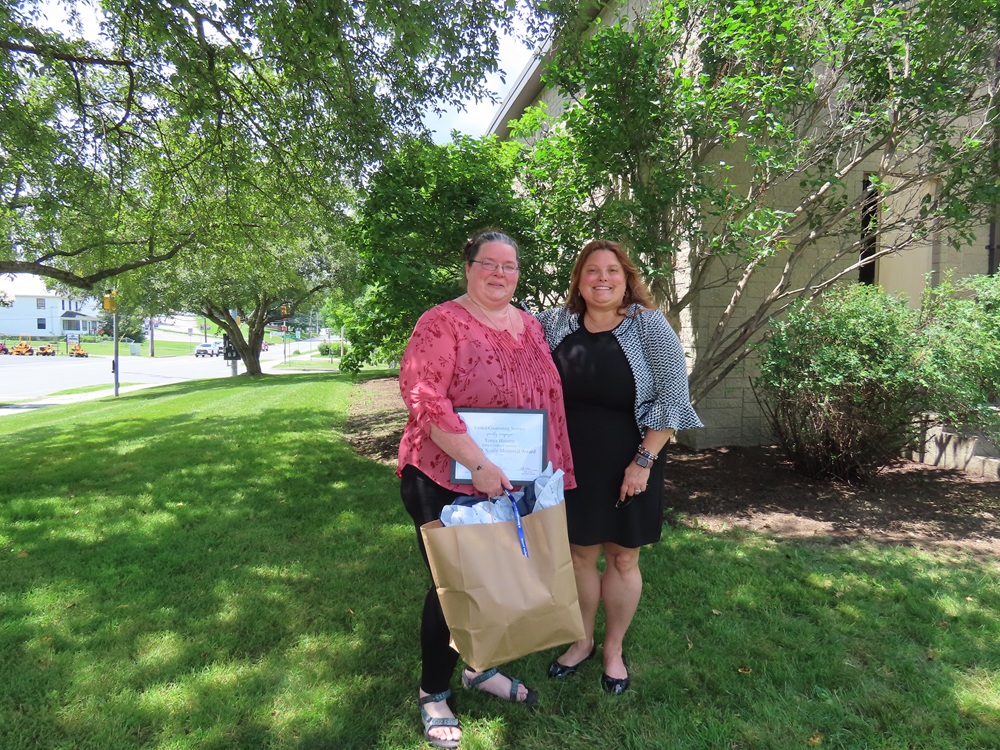 two women smile with a green, summer background behin them. Woman on the left holds a large-handled paper bag and a certificate.