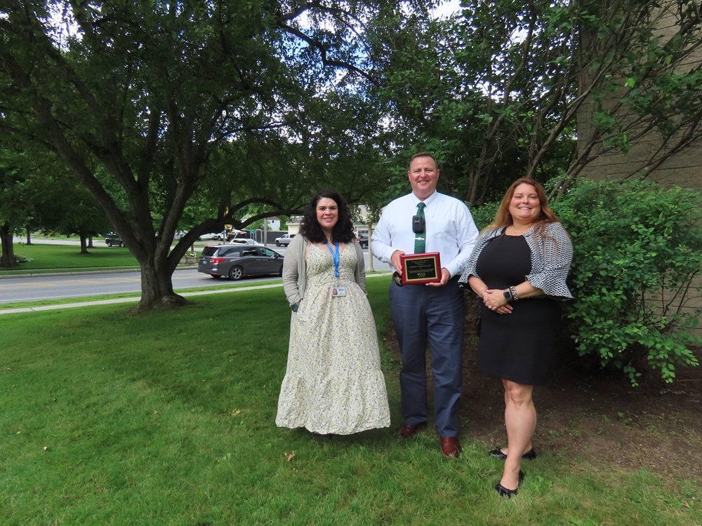 two women in dresses stand on either side of a man in a white button down shirt and a tie holding a plaque and smiling