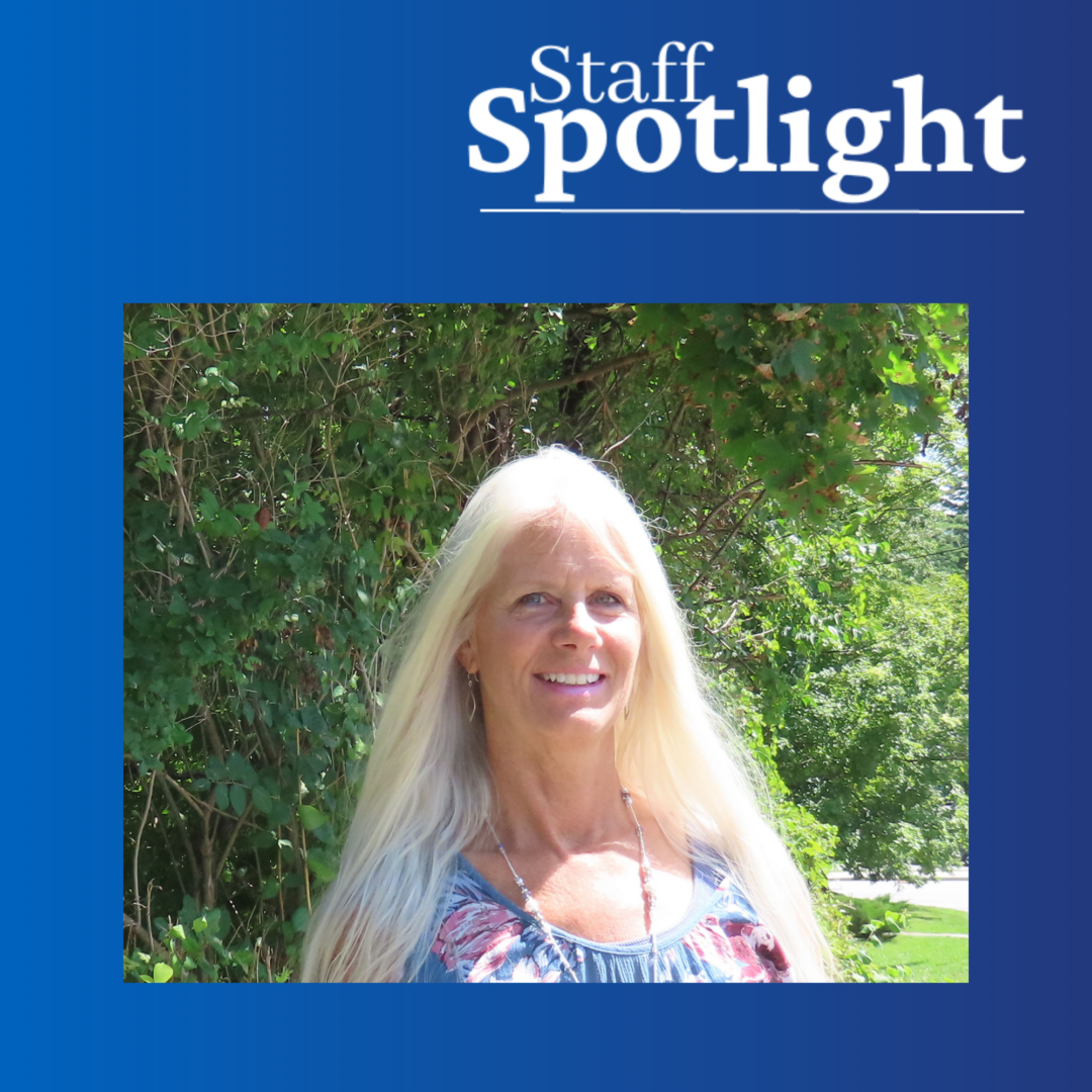 Woman with long silver hair wearing a floral blue and pink shirt smiles in the sunlight against a green leafy sunny background. Blue gradient background with 'Staff Spotlight' in top right of image.