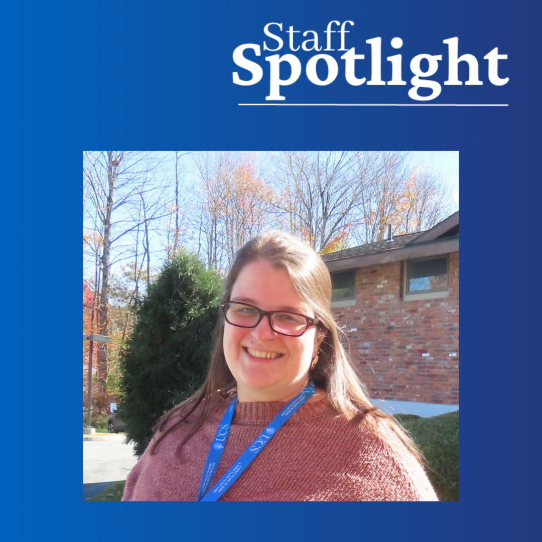 a woman with glasses and dark hair pulled back halfway wearing a blue lanyard and pink sweater stands outside of a brick building on a sunny day. Border graphic is blue and says 'Staff Spotlight'.
