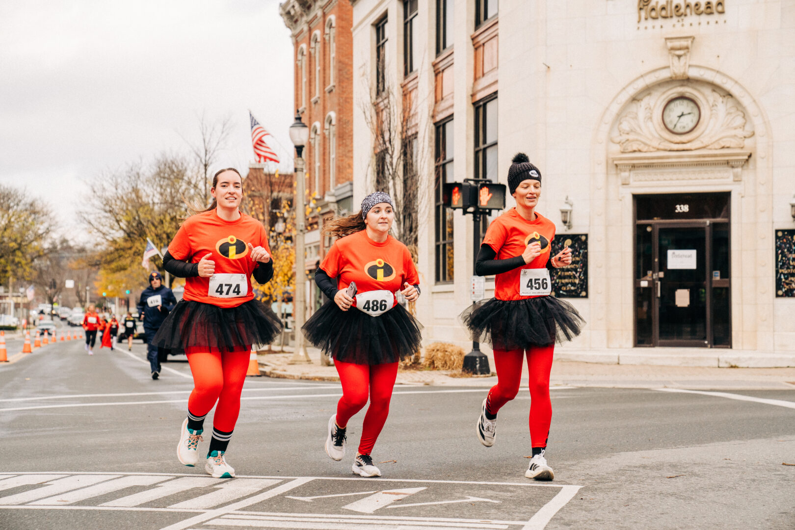 Three women in red superhero outfits with a red "i" on their t-shirts run across a historic-looking intersection.